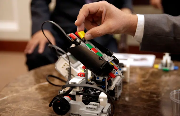 Uma mão a colocar uma peça num protótipo de um robot, pousado numa mesa, atrás vê-se a parte de baixo do tronco de uma pessoa, com uma mão na perna.